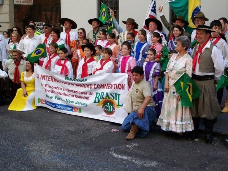 Grupo de pessoas com trajes tradicionais gaúchos em um desfile, segurando uma faixa do 4º Encontro Internacional do Tradicionalismo Gaúcho em New York/New Jersey