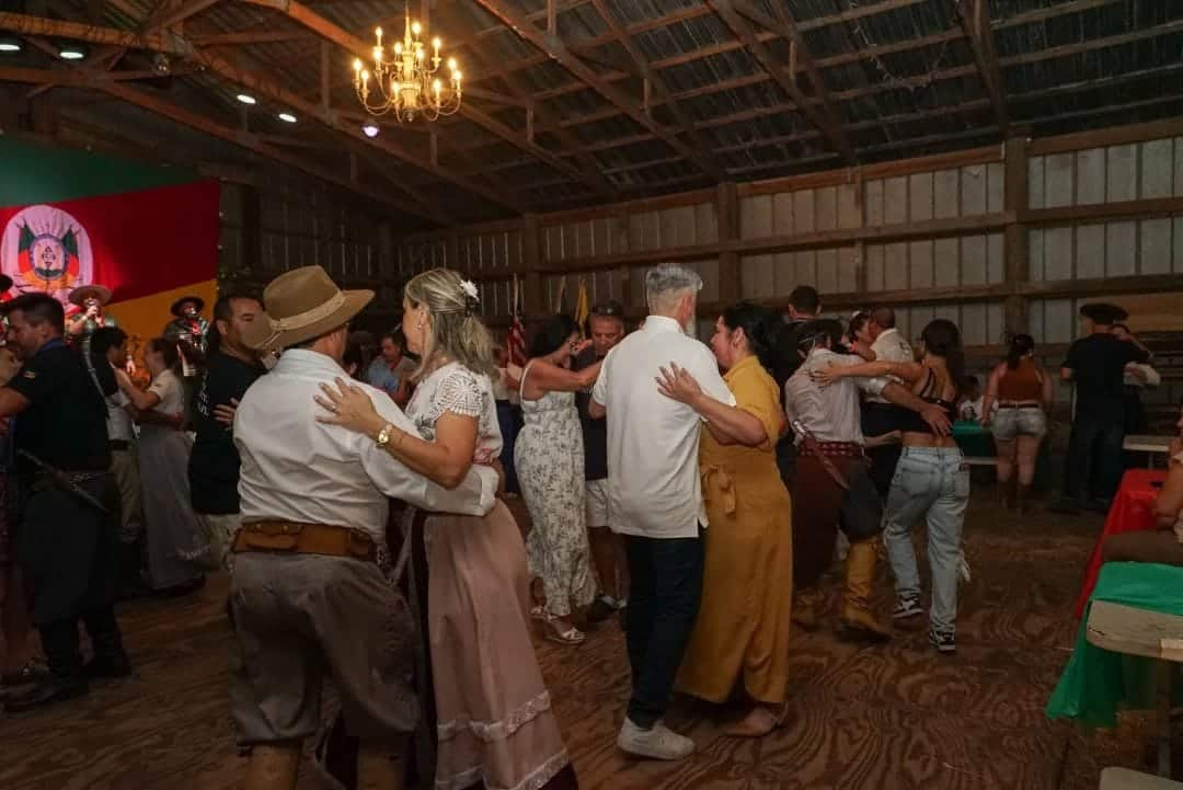 Casais dançando em um salão rústico durante uma festa, com palco e bandeira ao fundo