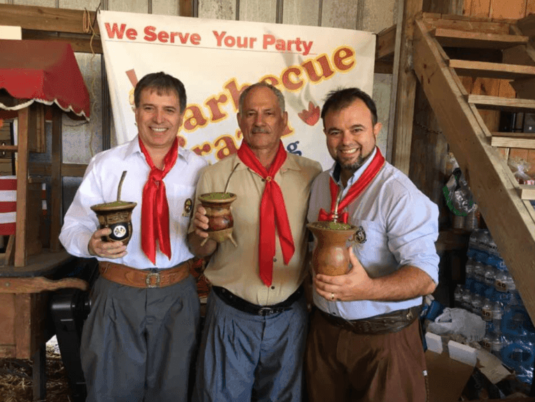 Três homens sorrindo, vestidos com lenços vermelhos, seguram cuias de chimarrão em frente a um banner de churrasco