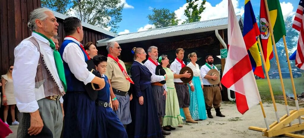 Grupo de pessoas vestidas com trajes tradicionais, em cerimônia ao ar livre, diante de várias bandeiras hasteadas