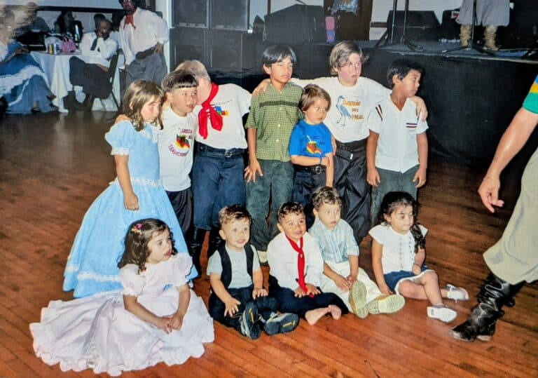 Grupo de crianças posando em uma festa ou apresentação, com roupas típicas e sentadas no chão de madeira