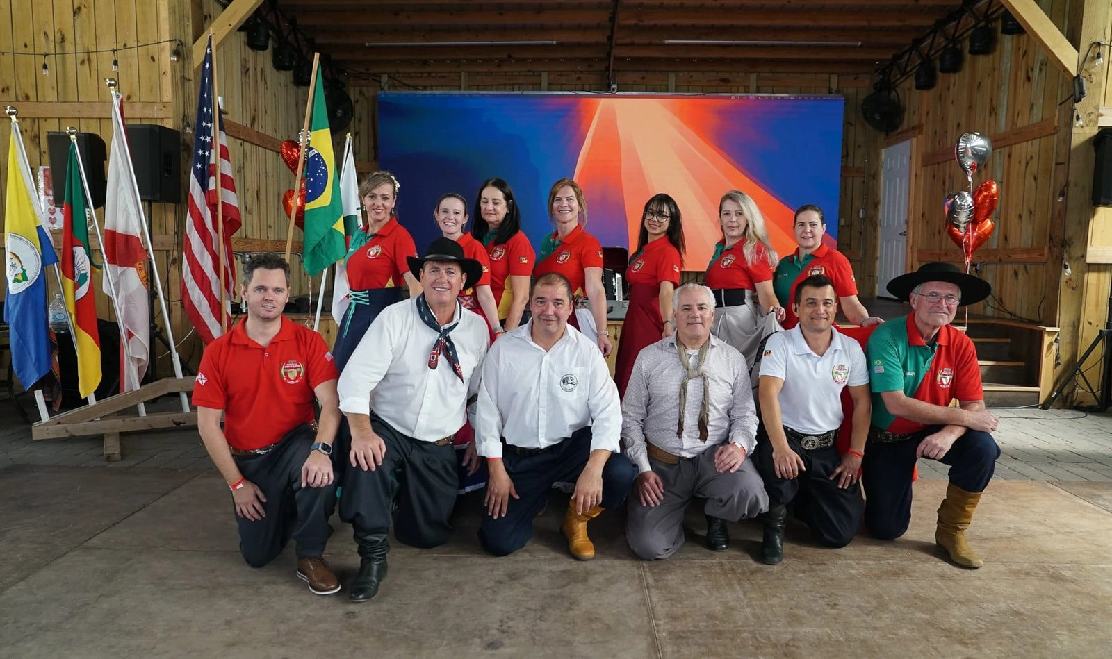 Grupo de pessoas posando em traje tradicional gaúcho, em um salão decorado com bandeiras e balões