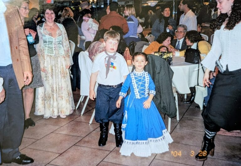 Crianças fantasiadas em uma festa, com adultos conversando ao fundo em um salão