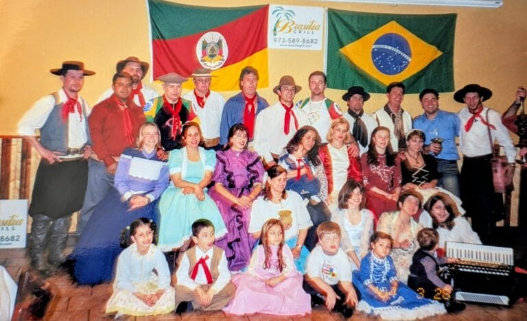 Grupo de pessoas vestidas com trajes gaúchos posando diante das bandeiras do Rio Grande do Sul e do Brasil