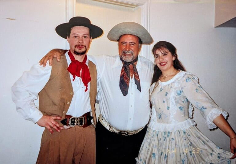 Três pessoas vestidas com trajes típicos de gaúcho posam sorrindo para a foto