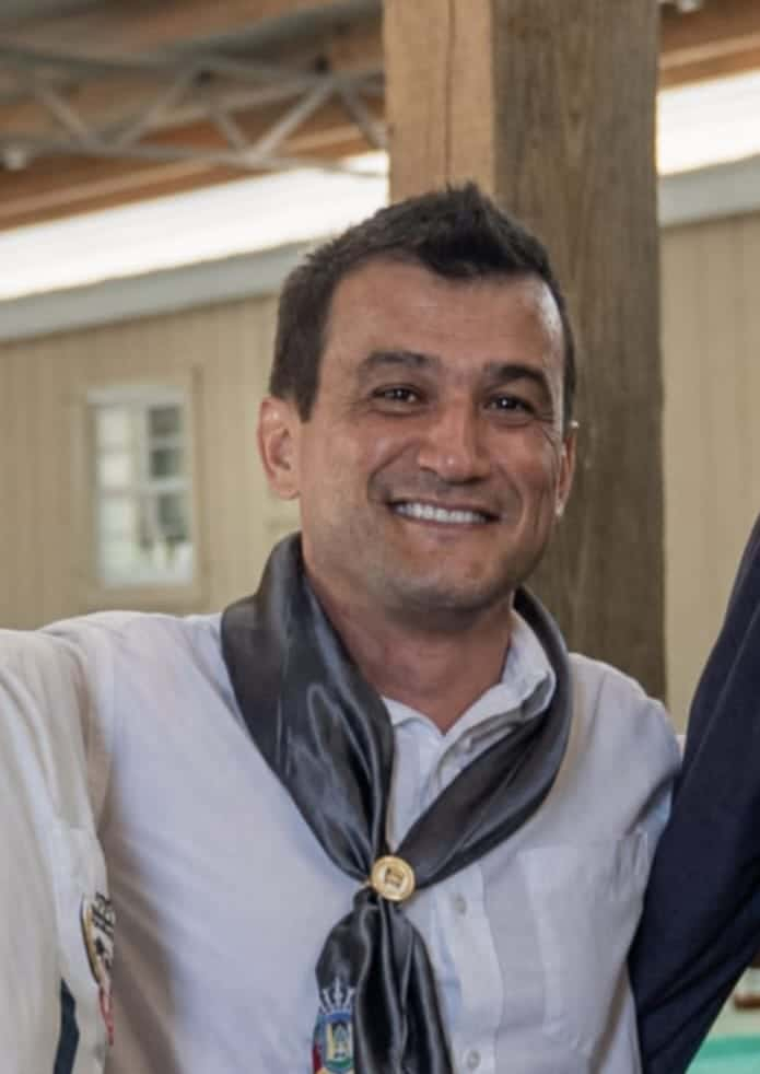 Homem sorrindo usando camisa branca e lenço preto no pescoço, em ambiente interno
