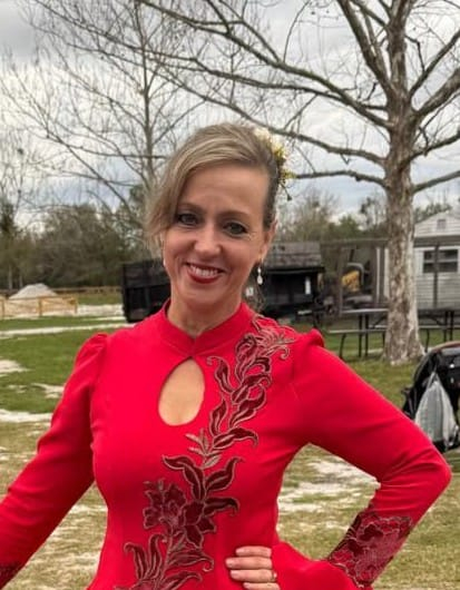 Mulher sorrindo de vestido vermelho bordado, em um ambiente externo com árvores ao fundo
