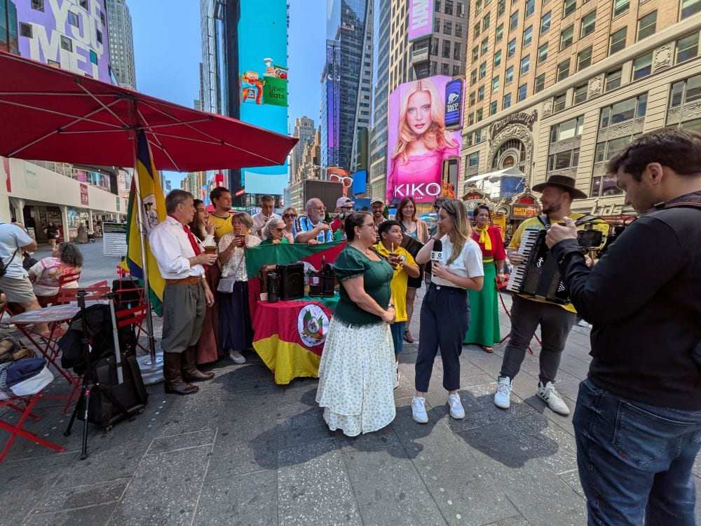 Grupo de pessoas gauchas reunidas na Times Square, com músicos tocando acordeão e uma repórter entrevistando participantes de uma celebração cultural de mateada 2026
