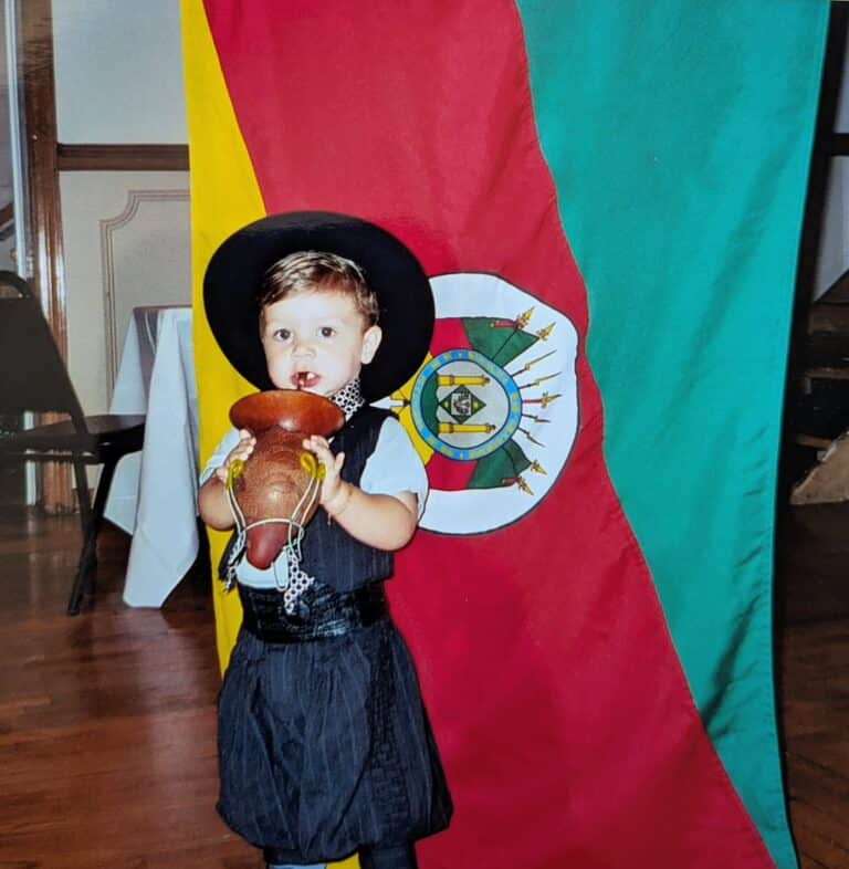 Criança vestida com traje tradicional segurando um instrumento musical, em frente à bandeira do Rio Grande do Sul