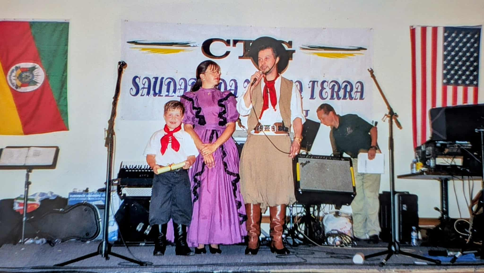 Grupo em palco durante apresentação musical, com adultos e uma criança vestidos de forma tradicional e microfone ao centro