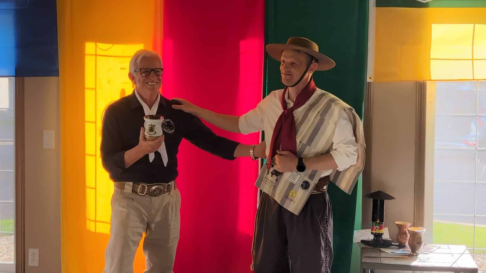 Dois participantes em trajes gaúchos diante de um painel nas cores amarelo, vermelho e verde, um deles segurando uma cuia.