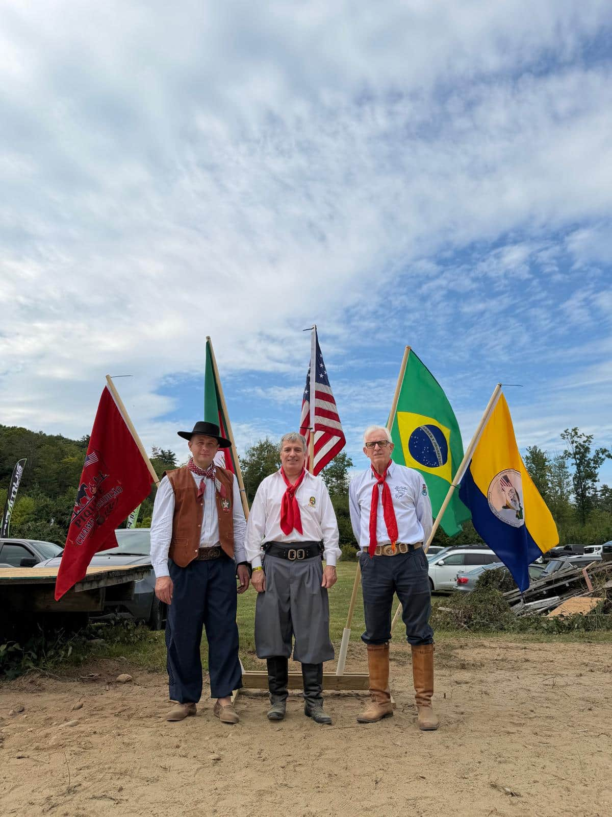 Três participantes em trajes gaúchos diante de bandeiras no evento de 30 de agosto de 2026 em New Hampshire.