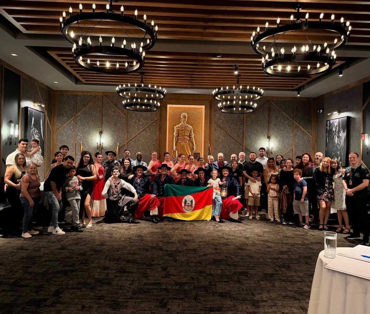 Grupo reunido em um salão, com participantes em trajes tradicionais gaúchos e a bandeira do Rio Grande do Sul.