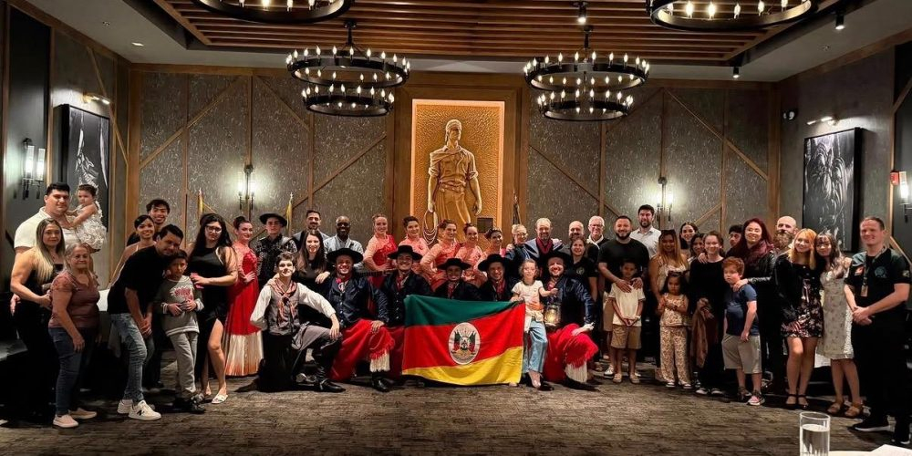 Grupo reunido em um salão, com participantes em trajes tradicionais gaúchos e a bandeira do Rio Grande do Sul.